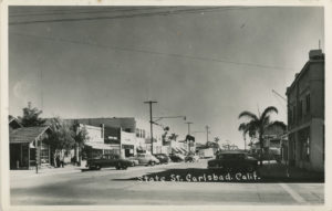 State Street, Carlsbad, California. Facing southeast from intersection of Grand Avenue. Two men are seated and one stands in front of 2906 State Street. Automobiles are parked along both sides of the street and a tanker truck can be seen at the Mobil gas station on the west side of the street. Captioned: State St. Carlsbad, Calif. (Courtesy of the Carlsbad City Library Carlsbad History Collection)